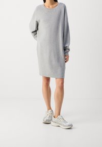 Robe grise en maille avec une coupe ample, un col large et des manches longues, associée à des chaussures de sport argentées avec lacets et un design texturé.