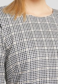 Primer plano de una persona rubia que lleva un top de patrón de houndstooth en blanco y negro con un escote redondo contra un fondo claro.