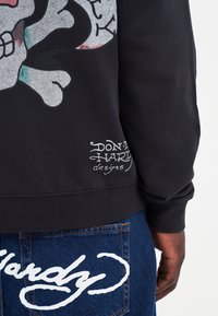 Felpa nera con una grande grafica di teschio e ossa incrociate, con una stampa testurizzata e logo bianco in basso a sinistra. Jeans in denim con cuciture visibili.
