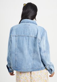 Giacca in denim azzurro chiaro con vestibilità rilassata, colletto a punte e due tasche laterali. Presenta cuciture in vista e un'etichetta rettangolare sulla parte posteriore.