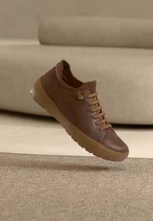 Baskets en cuir marron avec lacets et semelle couleur camel, flottant au-dessus d’un tapis beige texturé, avec un canapé doux en arrière-plan.