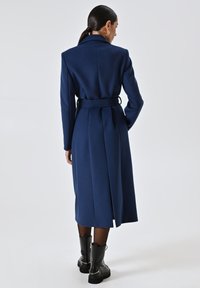 Langer marineblauer Mantel mit tailliertem Schnitt und Gürtelsaum, Rückenschlitzen, kombiniert mit schwarzen Knöchelstiefeln mit Kettenakzenten.
