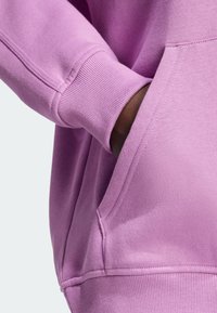 Tmavě hnědá ruka zasunutá do přední kapsy světle fialového mikinového svetru s vroubkovanými manžetami a lemem.