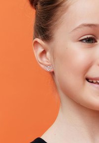 Srebrne kolczyki w kształcie serca z delikatnym otwartym wzorem, noszone na uchu młodej osoby na pomarańczowym tle.