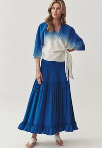 Top stile wrap in tessuto bianco e blu degradé, abbinato a una gonna blu a strati con orlo arricciato, indossato con sandali beige.
