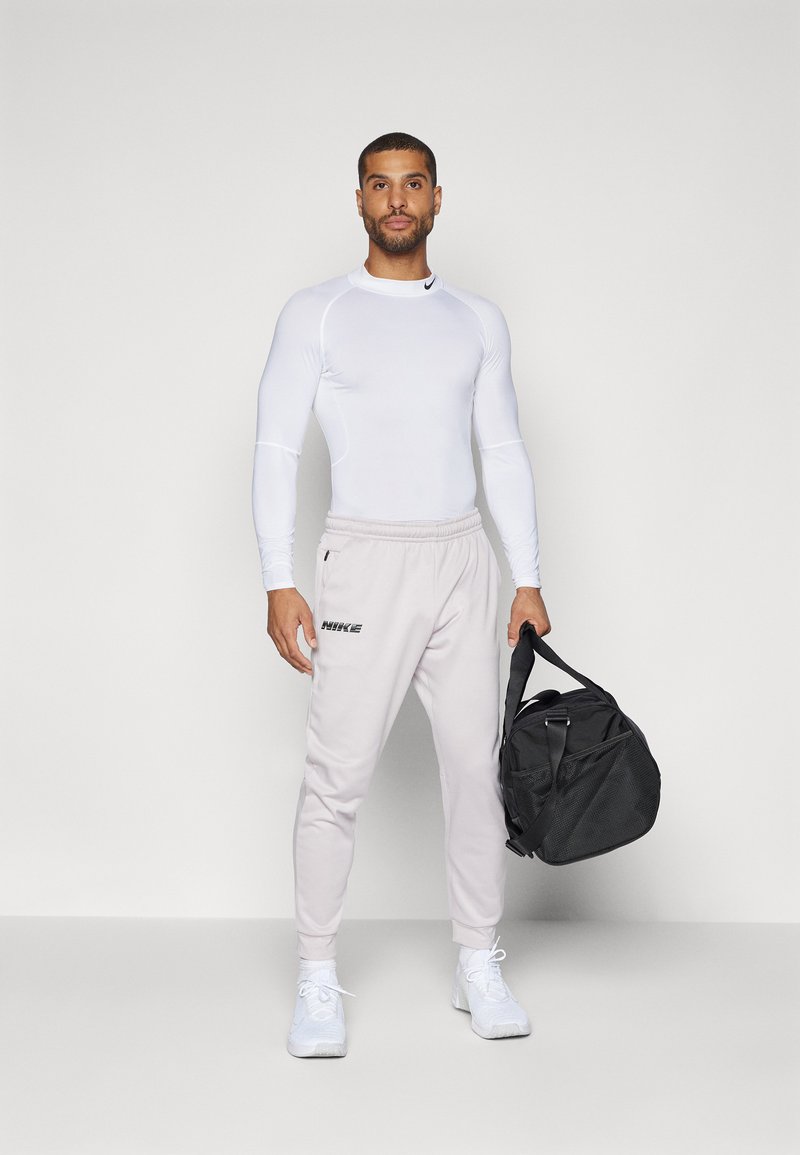 Haut de sport blanc à manches longues et joggeurs gris clair avec taille élastique ; logo Nike noir sur le pantalon. Tenant un sac de sport noir.