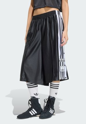 Osoba nosząca czarne szerokie szorty Adidas z białymi paskami po bokach, białe skarpetki Adidas i czarne czółenka Adidas na wysokim cholewce.