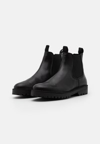 Une paire de bottines Chelsea en cuir noir avec des panneaux latéraux en élastique et des languettes de tirage, dotées de semelles à crans épaisses, présentées sur un fond blanc uni.