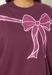 Kobieta ubrana w fioletową bluzkę z dużą, błyszczącą różową kokardą graficzną na klatce piersiowej i ramieniu.