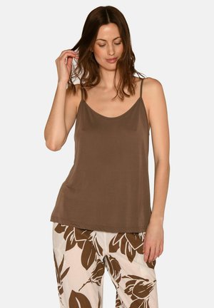 Kvinde iført en brun ærmeløs top og hvide bukser med store brune blomsterprint, stående mod en ensfarvet lys baggrund.