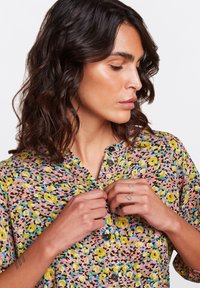 Camicia a maniche corte con bottone a fiori colorati su uno sfondo rosa, caratterizzata da motivi gialli e verdi, con colletto classico.