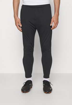 Man draagt zwarte atletische taps toelopende broek, witte sokken en zwarte sportschoenen met noppen, staand voor een effen achtergrond.