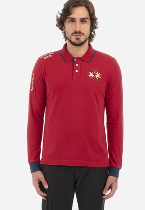 Šedý dlhý rukáv polo tričko s námorníckym golierom, vyšitým zlatým logom na hrudi a tmavomodrými manžetami. Obsahuje textové akcenty na pleciach.