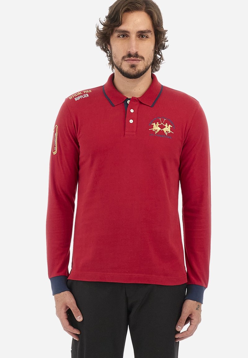 Rde majica s dolgimi rokavi polo stila z mornarsko modrim ovratnikom, vezenem zlatim logotipom na prsih in temno modrimi manšetami. Ima tudi akcentne napise na ramenih.
