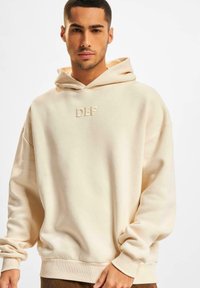 Hoodie couleur crème avec une poche kangourou, des poignets côtelés et une coupe décontractée. Présente un logo "DEF" en relief sur la poitrine.