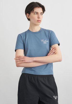 Persoon met kort donker haar die een blauw Carhartt Wip T-shirt en zwarte broek draagt, met gekruiste armen, kijkend naar de zijkant.