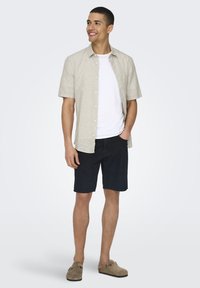 Camisa de manga corta color beige claro sobre una camiseta blanca, combinada con pantalones cortos azul oscuro y zapatos marrones sin cordones; diseño sencillo y ajuste casual.