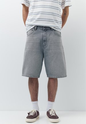 Mand iført grå denimshorts, hvide mellemlange sokker, mørke sneakers og en hvid t-shirt med striber, stående mod en ensartet lys baggrund.