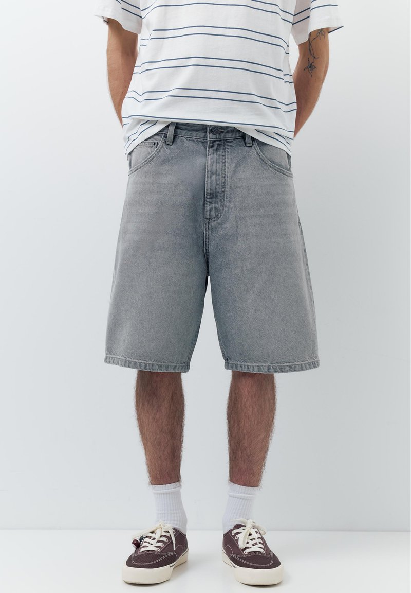 Homme portant un short en jean gris, des chaussettes blanches, des baskets foncées et un t-shirt blanc à rayures, debout devant un fond clair uni.