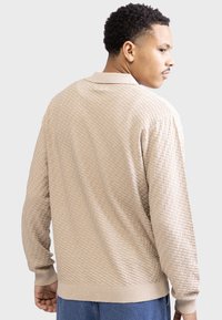 Pullover longo bege texturizado com um padrão geométrico subtil, apresentando um colarinho clássico e punhos e bainha canelados.