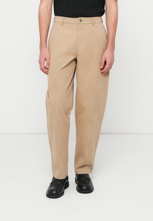 Άνδρας που φοράει μπεζ παντελόνι με ίσια γραμμή και πλαϊνές τσέπες, καθώς και μαύρα δερμάτινα παπούτσια τύπου slip-on, στέκεται μπροστά σε λευκό φόντο.