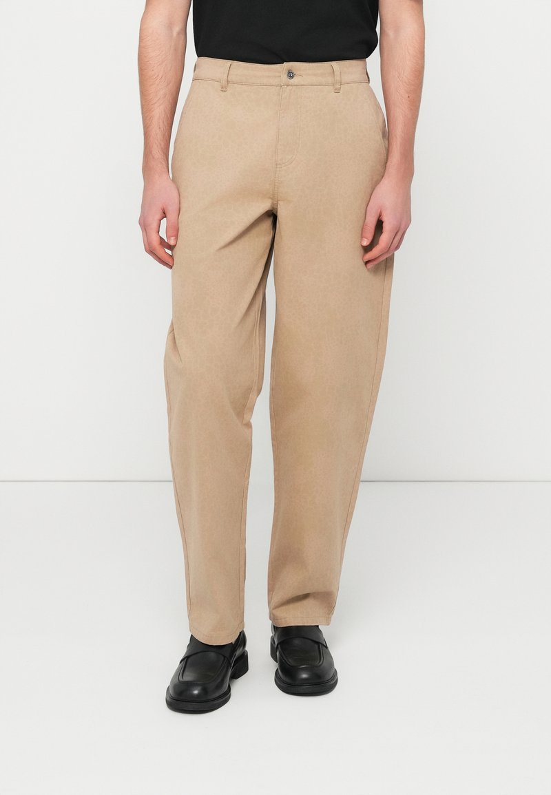 Άνδρας που φοράει μπεζ παντελόνι με ίσια γραμμή και πλαϊνές τσέπες, καθώς και μαύρα δερμάτινα παπούτσια τύπου slip-on, στέκεται μπροστά σε λευκό φόντο.