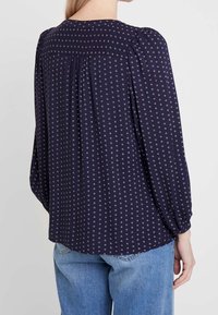 Blusa navy con maniche lunghe a sbuffo, caratterizzata da un piccolo motivo floreale bianco. Pieghe sulla schiena per aggiungere volume; tessuto morbido con una texture liscia.