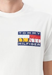 Witte katoenen t-shirt met een gekleurd rechthoekig logo met "TOMMY HILFIGER", "85" en "NYC" in verschillende vette lettertypen. Korte mouwen.