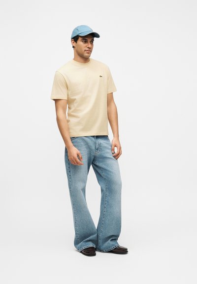 Homme portant une chemise beige à manches courtes, un jean bleu clair à jambes larges, des chaussures foncées et une casquette de base-ball bleu clair, debout devant un fond uni.