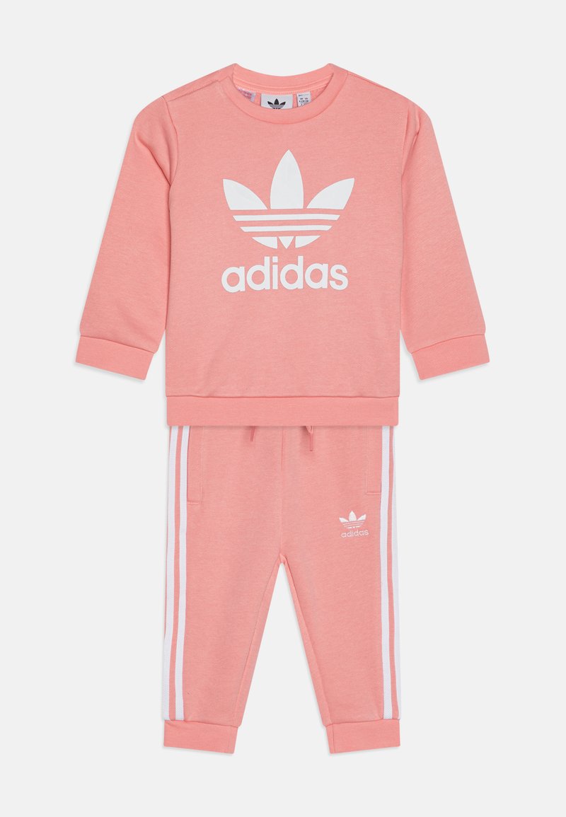 Różowy bawełniany dres z topem o okrągłym dekolcie z białym logo Adidas oraz trzema białymi paskami na spodniach. Zawiera ściągacze u dołu rękawów i w pasie.