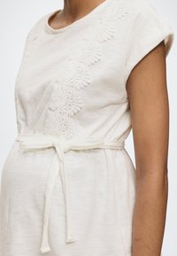 Persoană însărcinată, purtând o rochie crem cu mâneci scurte, detalii din dantelă și curea din material textil legată, prezentată de la umăr până la șold.