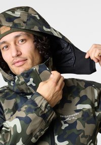 Camouflage-takki hupulla, jossa on vaihtelevaa vihreän ja mustan sävyä. Sisältää mustan sisävuoren, nepparit ja logon yksityiskohtana.