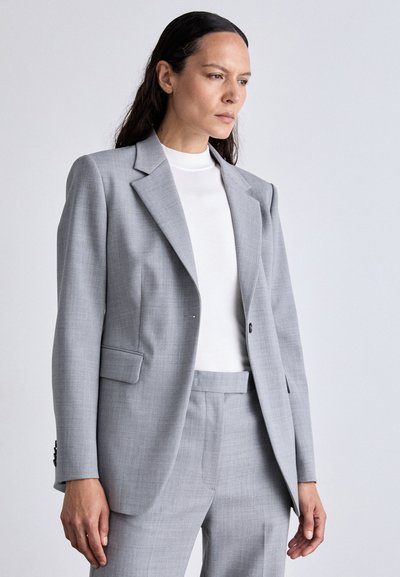 Blazer cinza claro com um corte ajustado, lapelas de pico e bolsos laterais, combinado com um top de gola alta branca e calças a condizer.