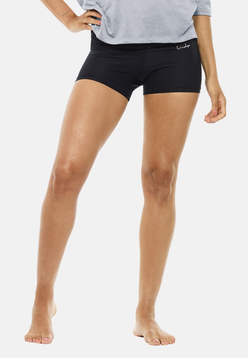 Black athletic shorts with a smooth texture, featuring a wide waistband. The model stands barefoot, wearing a loose gray top.
