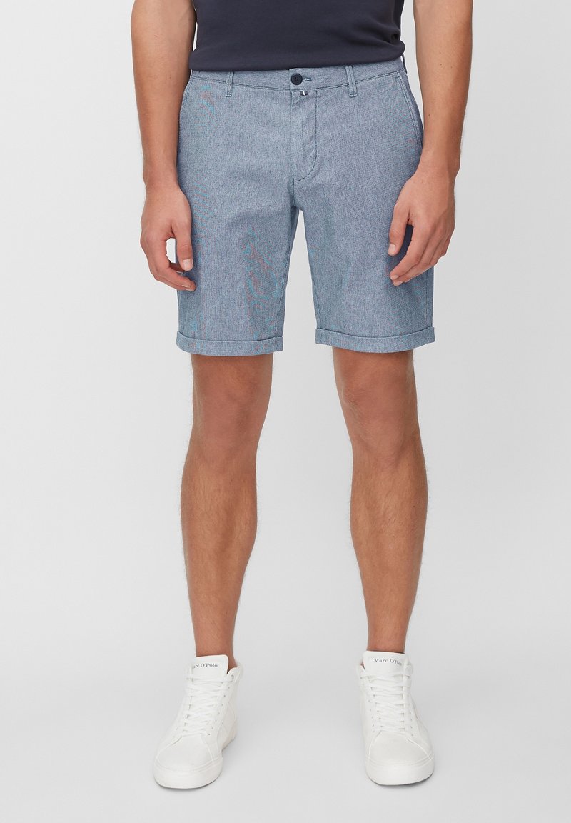 Homme portant un short bleu clair avec ourlet retroussé, un t-shirt noir et des baskets blanches à lacets, debout devant un fond uni clair.