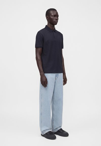 Homme debout portant un polo noir à manches courtes, un jean large bleu clair et des baskets noires sur un fond clair uni.
