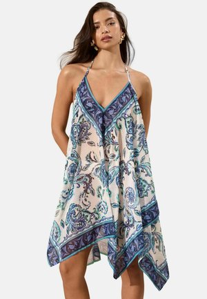 Femme portant une robe dos nu ample avec des motifs floraux bleus et verts et des motifs paisley sur un fond clair.