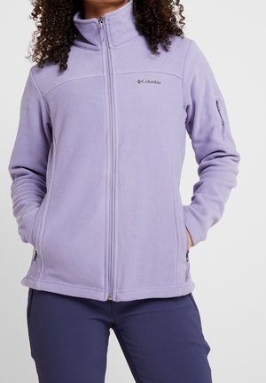 Femme portant une veste polaire Columbia violet clair avec une fermeture éclair complète et les mains dans les poches, associée à un pantalon foncé.