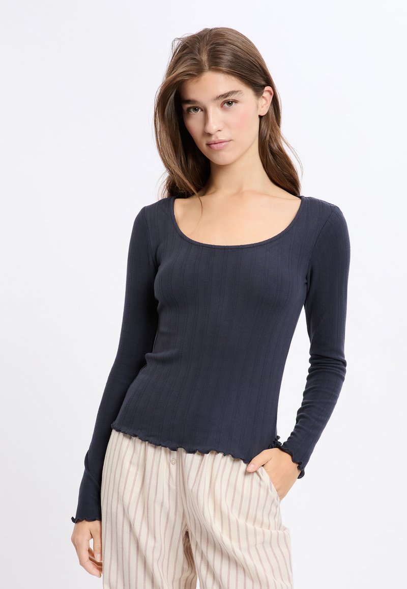 Długi rękaw, granatowa bluzka z prążkowanej tkaniny z dekoltem w serek. Posiada falbaniasty dół i dopasowany krój. Połączona z jasnymi spodniami w paski.