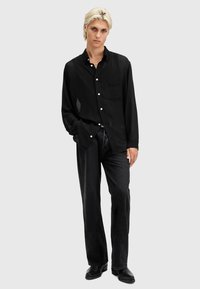 Cămașă neagră cu mânecă lungă, tip button-up, dintr-un material texturat, având un buzunar frontal și contraste subtile de model, asortată cu o pereche de blugi negri, largi.
