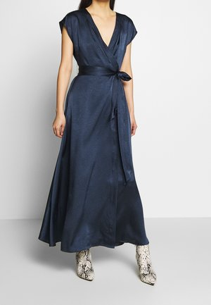 Woman wearing a navy blue wrap dress with a tied waist belt and white snakeskin-patterned ankle boots, standing against a plain background.