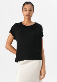 Top nera a maniche corte con scollatura drappeggiata, vestibilità rilassata e tessuto morbido. Abbinato a una gonna di tessuto liscio e chiaro.