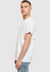 Weißes Baumwoll-T-Shirt mit kurzen Ärmeln, Rundhalsausschnitt und rotem gesticktem Text auf der Brust. Getragen mit blauen Jeans.