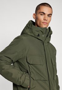Olívazöld kabát kapucnival. Elöl cipzárral, két nagy mellzsebbel és állítható húzózsinórokkal a tökéletes illeszkedésért. Simább, vízálló anyagból készült.
