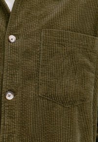 Chemise en velours côtelé vert olive avec texture à côtes verticales, fermeture à boutons et une poche poitrine unique avec une finition soignée.