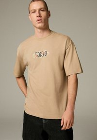 Camiseta de manga corta beige, hecha de algodón, con cuello redondo y un gráfico multicolor de "TOKYO" en el pecho.