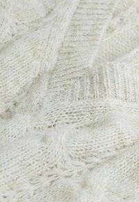 Tissu en tricot couleur crème avec une texture douce et duveteuse. Présente un motif en câble et des accents côtelés, offrant une profondeur visuelle et de la chaleur.