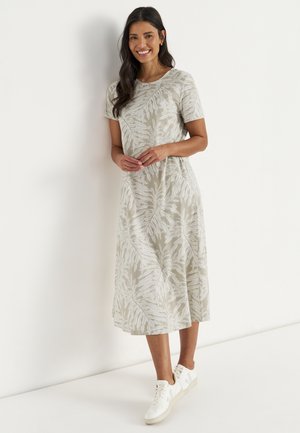 Kvinde står smilende, iført en beige midi-kjole med hvide bladmønstre og hvide sneakers mod en ensfarvet hvid baggrund.