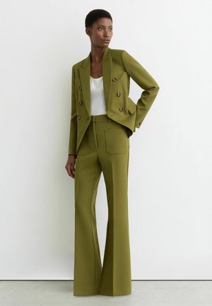 Donna che indossa un blazer doppiopetto verde oliva e pantaloni abbinati a vita alta e gamba larga, in piedi contro un muro bianco semplice.