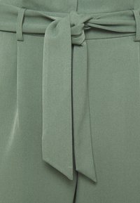 Tissu vert clair avec une texture lisse, présentant une ceinture nouée en nœud. La ceinture a une largeur uniforme et complète le pantalon en dessous.